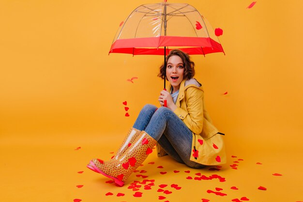 Эмоциональная молодая женщина в желтых резиновых туфлях, сидя на полу с бумажными сердечками. Внутреннее фото вдохновленной кудрявой девушки, позирующей с милым зонтиком.