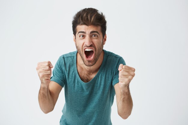 Emotions and achievement concept. Close up shot of happy successful casually weared student or employee screaming with winning expression, fists pumped, celebrating success on a white wall