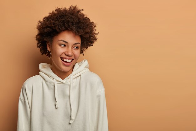 Emotions and lifestyle concept. Happy delighted dark skinned curly woman wears white sweatshirt, laughs and has fun, looks aside