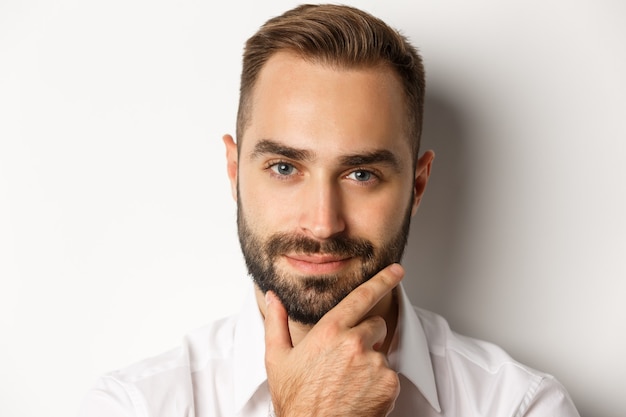 Foto gratuita concetto di emozioni e persone. primo piano del volto dell'uomo premuroso bello sorridente soddisfatto, toccando la barba e pensando, in piedi