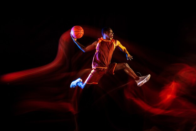 Emotions of winner. African-american young basketball player of red team in action and neon lights over dark studio background. Concept of sport, movement, energy and dynamic, healthy lifestyle.