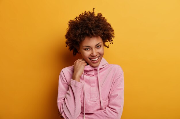 Emotions, youth and people concept. Amused pretty woman laughs sincerely, has shy expression, has interesting idea, keeps hand on hoodie, enjoys life, poses indoor against yellow wall