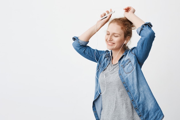 Emotive adorable teenage ginger girl smiling while listening to music via earphones, holding smartphone and lifting hands in dance