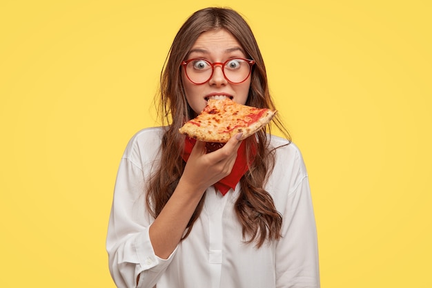 Emotive beautiful lady bites delicious pizza, looks directly has time for snack, visits pizzeria, surprised with low prices, models over yellow wall. people, fast food and nutrition