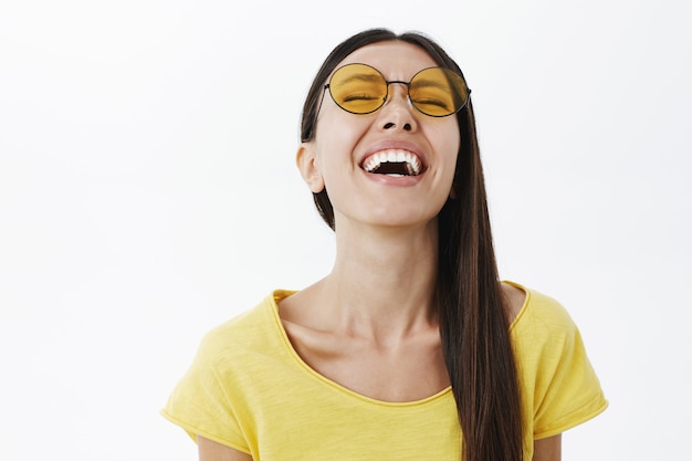 Foto gratuita emotiva donna attraente spensierata e ottimista con i capelli scuri in occhiali da sole gialli rotondi alzando la testa mentre ride