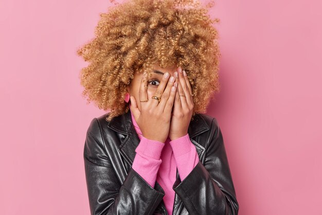 Emotive cheerful young woman covers eyes with palms peeks through fingers tries to hide dressed in leather jacket isolated over pink background tries to look at something. Happy emotions concept