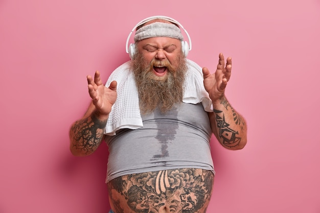 Emotive chubby hipster listens music in headphones, sings song loudly, dressed in sports wear, has fitness training to loose weight, poses against rosy wall. Athlete thick bearded man indoor