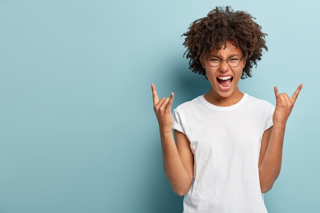 Emotive dark skinned woman makes rock n roll sign, says I will rock this party, yells loudly, wears round spectacles