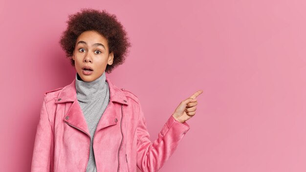 Emotive female teenager with Afro hair keeps jaw dropped from shock demonstrates amazing promo, pointing at copy space on pink wall