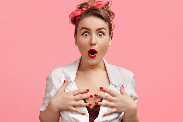 Emotive gorgeous lady keeps hands on breast, stares with bugged eyes and bated breath, can't believe in something amazing, wears red headband, has red painted lips and manicure, isolated on pink