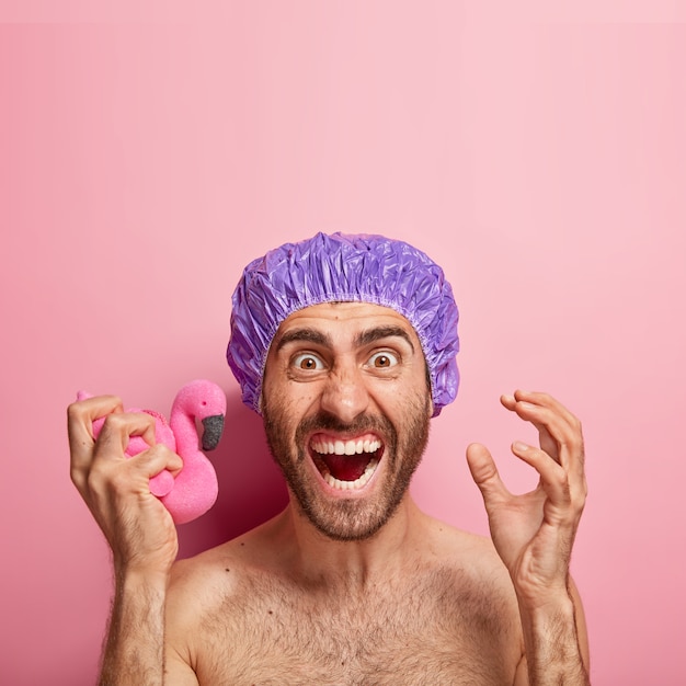 감정적 인 남자는 적극적으로 몸짓으로 짜증이 나며 부드러운 분홍색 플라밍고 스폰지를 들고 샤워 캡을 착용합니다.
