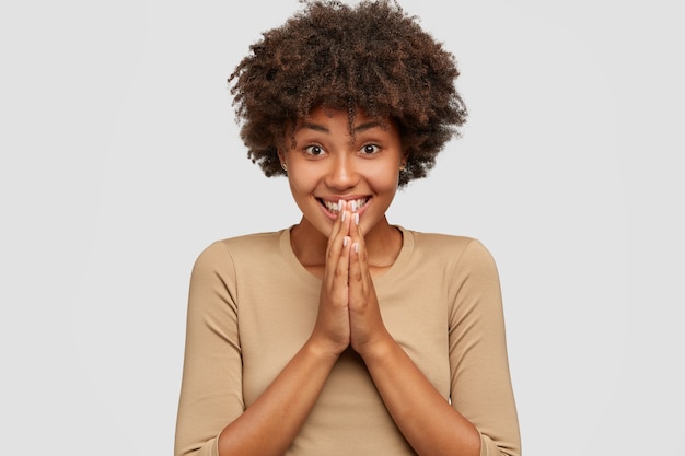 Emotive pleased dark skinned girl has toothy smile, keeps hands in praying gesture