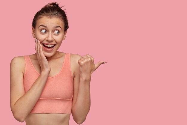  emotive pleased sportswoman looks gladfully aside, touches cheek with hand, points with thumb aside, wears casual top, isolated over pink wall with copy space for advertisement