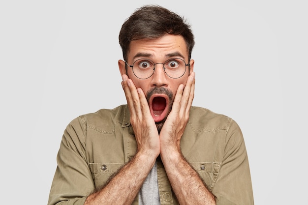 Emotive scared shocked male with widely opened mouth hears impressive rumor, stares with stunned expression, being in stupor, wears round glasses, isolated over white wall