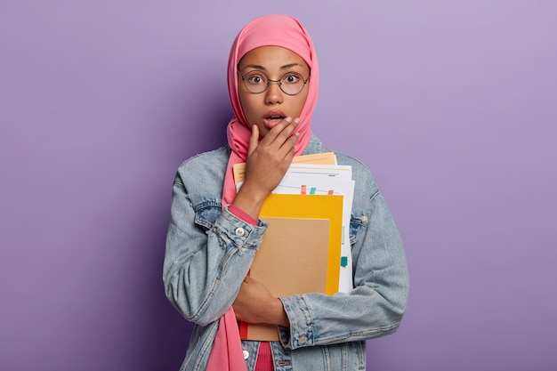 Emotive stupefied dark skinned muslim woman in hijab opens mouth from amazement, holds some papers and notepads