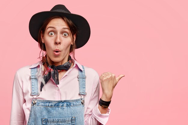 Emotive stupefied young European female farmer in casual overalls and hat, indicates with thumb aside