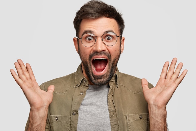 Free photo emotive surprised amazed happy male clasps hands, opens mouth opened and eyes popped out, can`t believe in great success, wears round spectacles, dressed in fashionable shirt, isolated over white wall