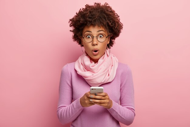 Emotive surprised dark skinned Afro female blogger makes publicaion on mobile phone