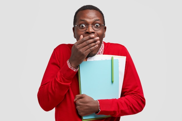 Emotive surprised dark skinned young man covers mouth with palm, has joyful expression, recieves excellent mark on exam, carries textbook, papers closely