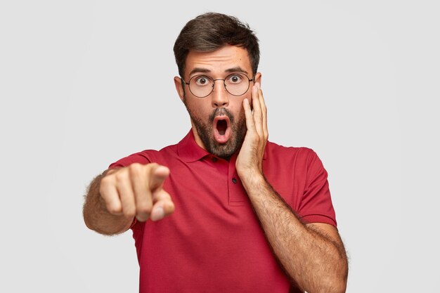 Emotive terrified Caucasian male with dropped jaw, notices somethning terrible on street, points into distance with index finger, dressed in red t-shirt, isolated on white wall. Reaction concept