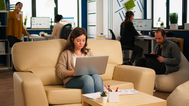 Сотрудник держит ноутбук, сидя в зоне отдыха на удобном диване, печатая на компьютере, улыбаясь, в то время как различные коллеги работают в фоновом режиме. Многонациональные коллеги планируют новый финансовый проект в компании