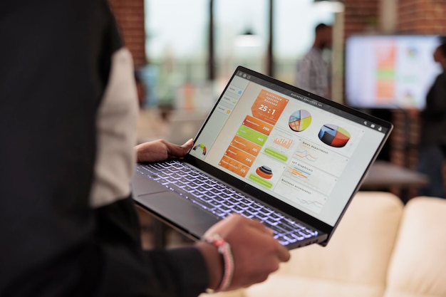 Employee holding laptop with business diagrams in company office, management presentation of diagrams and graphs on computer screen. Data report and research information on pc. Close up.