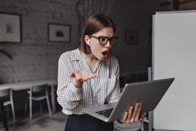 Сотрудник смотрит на открытый ноутбук с удивлением и разочарованием. Портрет деловой женщины в очках и наушниках в белом офисе.