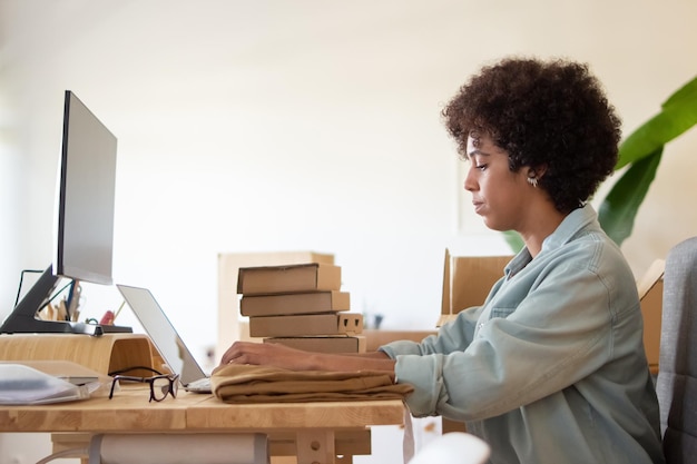 Free photo employee of online shop using laptop. serious african american businesswoman sitting at tab;e with stack of packages. logistics concept