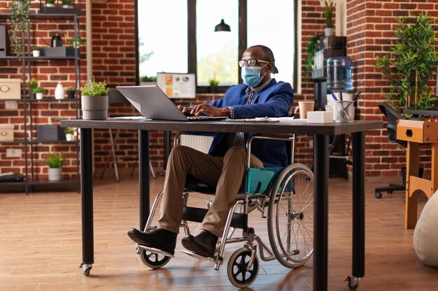 Сотрудник с ограниченными возможностями в маске для лица и работает над бизнес-проектом. Предприниматель, сидящий в инвалидной коляске, выполняет финансовую и маркетинговую работу во время пандемии коронавируса. Человек на работе