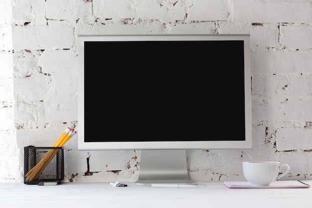 Foto gratuita schermo monitor nero vuoto su muro di mattoni bianchi con matite, note e tazza di caffè. copyspace,