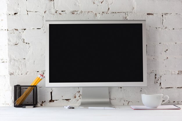 empty black monitor screen on white brick wall with pencils, notes and cup of coffee. Copyspace,