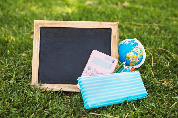 Empty chalkboard near stationery and globe on grass