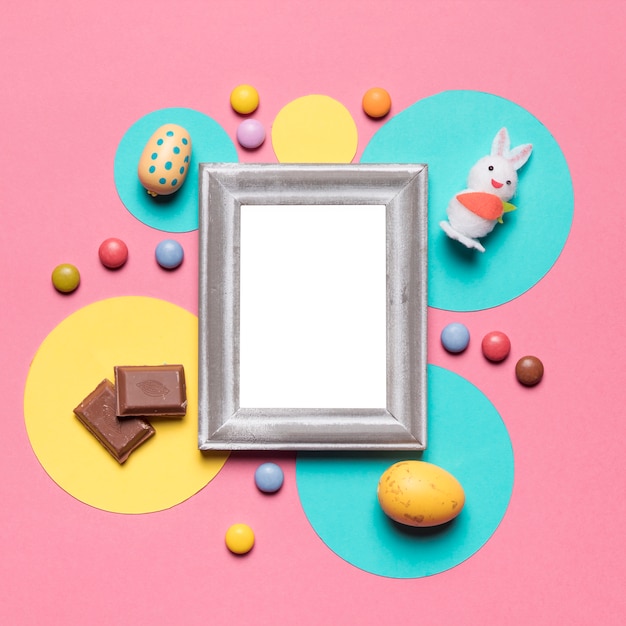 An empty frame surrounded with easter eggs; bunny; candies and chocolate pieces on pink backdrop