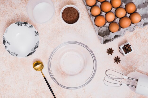An empty glass bowl; plate; flour; cocoa powder; eggs carton; star anise and electric mixer on kitchen counter