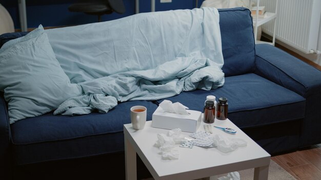 Empty living room with medicine against virus infection on table. Nobody in space with medicaments, drugs, capsules, bottles of pills and thermometer. Cup of tea and tissues for person with flu