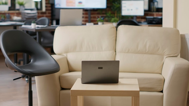 Empty startup office with couch and laptop computer, place decorated with business supplies and instruments. Nobody in company workspace to work on strategy and do financial presentation.