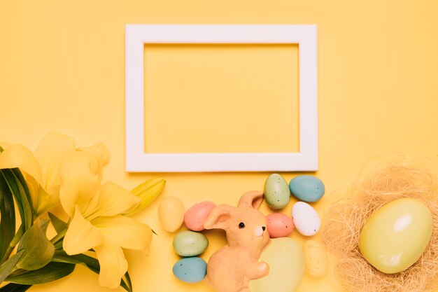 An empty white border frame decorated with lily flowers; rabbit figurine and easter eggs on yellow background