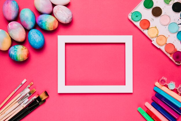 An empty white border frame with easter eggs; paint brushes; felt tip pens and water color paint box on pink background