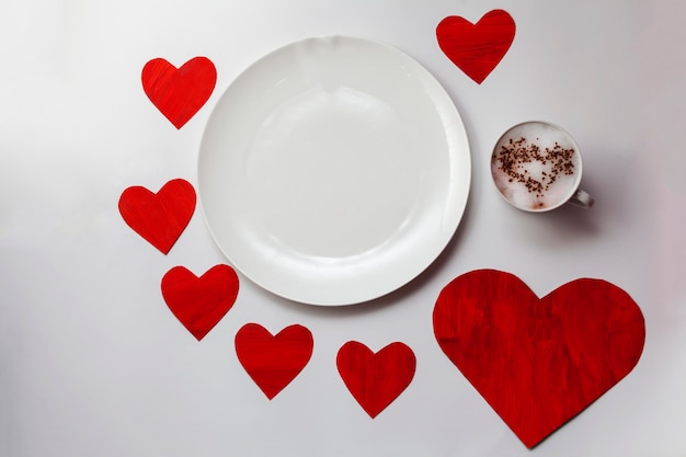 Пустая белая тарелка на стол с красными сердцами вокруг и чашка с горячим напитком и окрашенные сердца на пену.