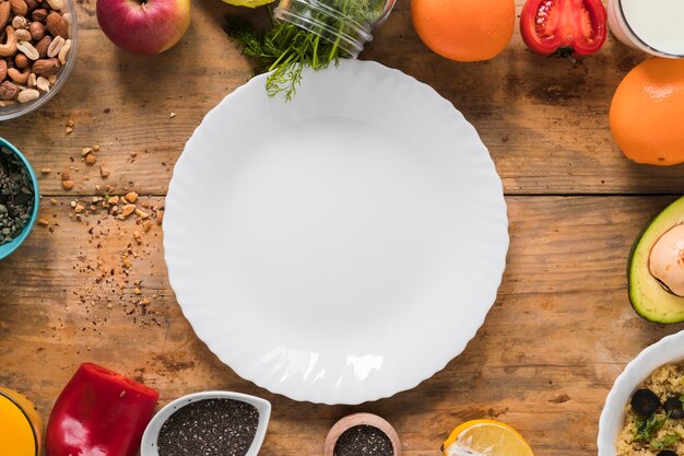 Empty white plate surrounded by dryfruits; vegetables; fruits on wooden table