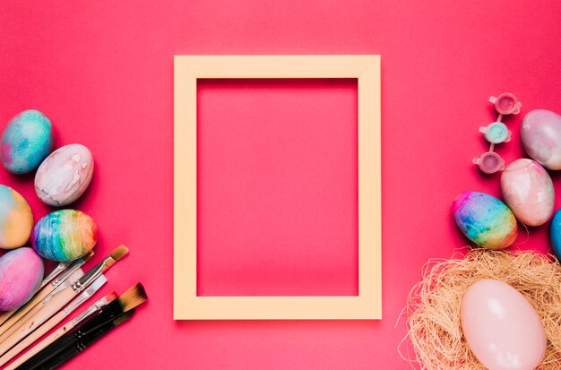 An empty yellow border frame with colorful easter eggs and paint brushes on pink backdrop