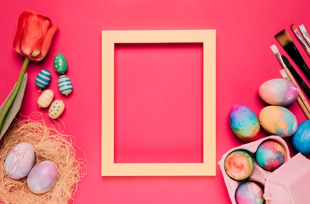 An empty yellow border frame with colorful easter eggs; paint brushes; tulip on pink backdrop