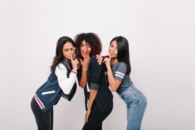 Enchanting african girl with red manicure cover mouth with hand while posing beside university mates. Fascinating latin young woman with shiny hair having fun with friends and laughing.