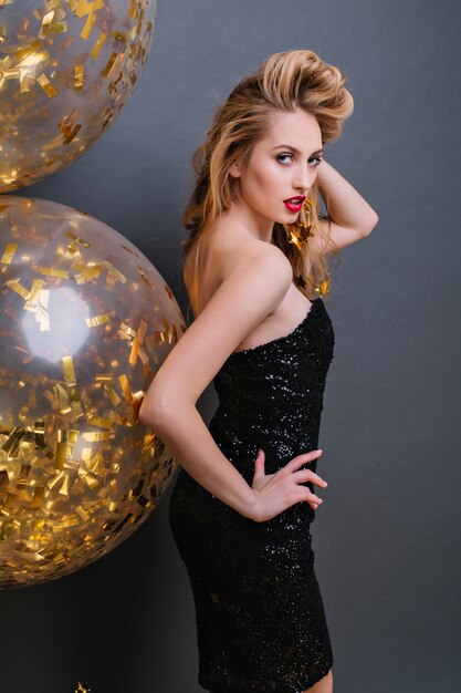 Enchanting blonde girl looking over shoulder and plays with her hair during party photoshoot. Slim fair-haired lady wears black dress posing with golden balloons.