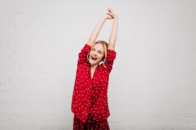 Enchanting blonde woman in red sleepwear stretching on light wall with smile. Cute european girl in pajamas enjoying good morning and laughing.