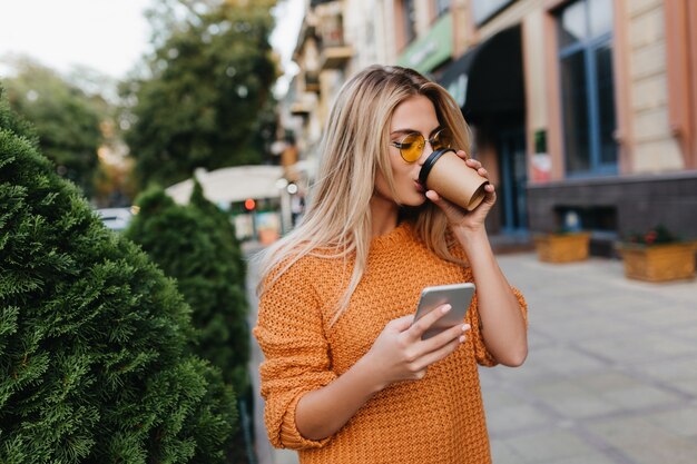 路上でコーヒーを飲みながら電話メッセージを待っている魅惑的な金髪の若い女性