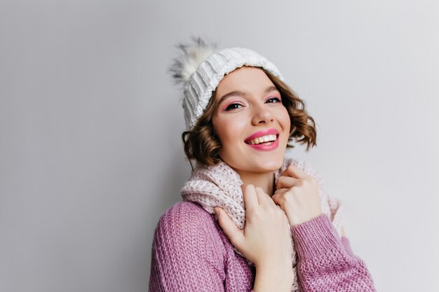 보라색 스웨터와 흰색 액세서리에 포즈를 취하는 행복 한 얼굴 표정으로 매혹적인 곱슬 소녀. 아름다운 백인 여성 모델의 실내 사진은 니트 소프트 모자와 스카프를 착용합니다.