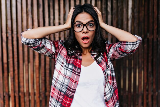 Enchanting dark-eyed girl expressing surprised emotions in spring day. Outdoor shot of shocked stylish woman isolated on wooden wall.