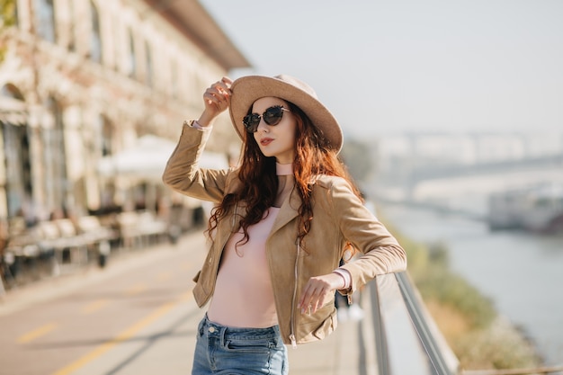 Enchanting ginger woman touching her hat while posing on sea wall