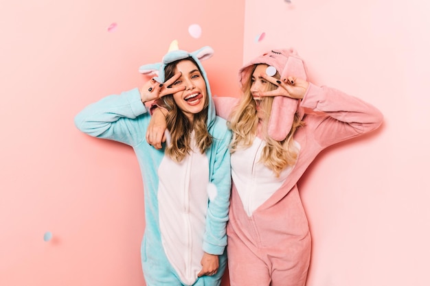 Enchanting girl in pink kigurumi posing with peace sign Studio shot of charming female model wears funny pajamas